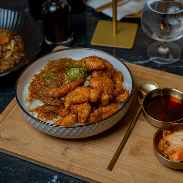 Korean fried chicken and japchae noodles over rice, with kimchi and savory pancakes on a wooden board.