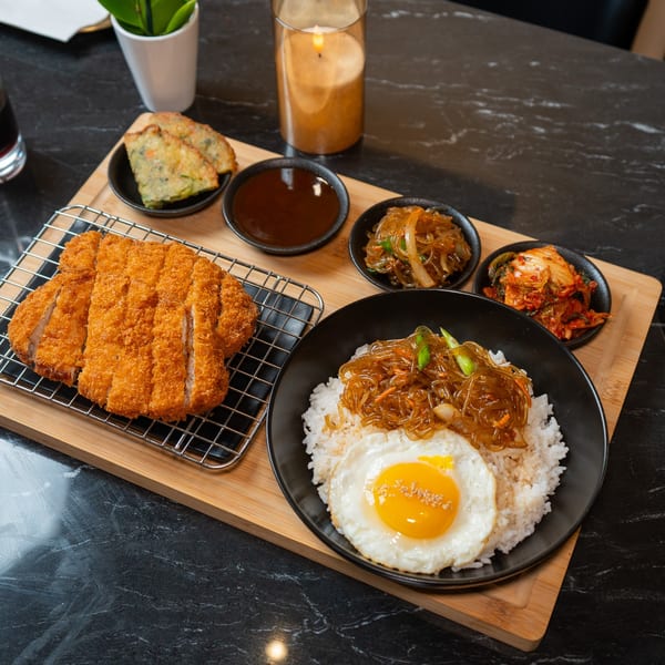 Korean meal with crispy fried pork cutlets, rice, fried egg, japchae, kimchi, and dipping sauces.