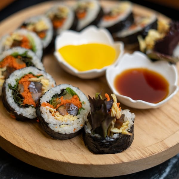 Sliced kimbap with various fillings, served on a wooden tray with two dipping sauces.