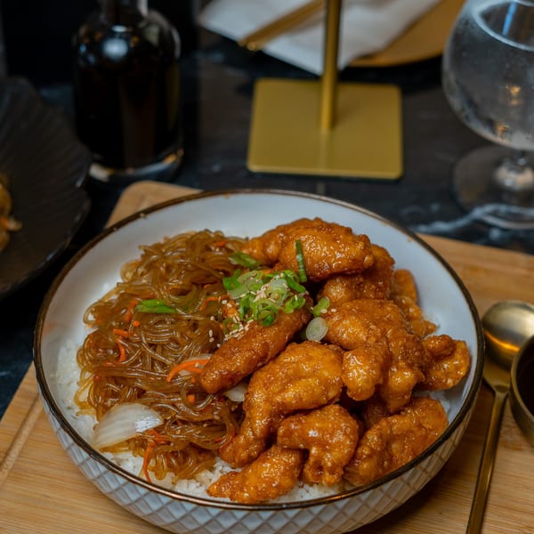 A bowl of Korean fried chicken and japchae noodles over rice on a wooden board.