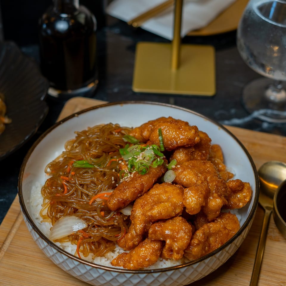 A bowl of Korean fried chicken and japchae noodles over rice on a wooden board.