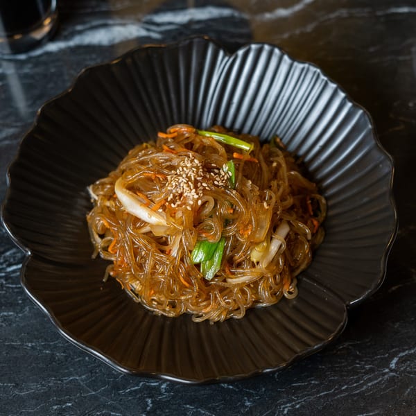 Japchae, Korean glass noodles with vegetables and sesame seeds, served in a black bowl on a marble table.