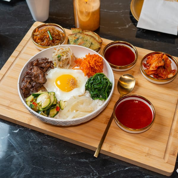 A colorful bibimbap bowl with beef, egg, and vegetables, served with kimchi and sauces on a wooden board.