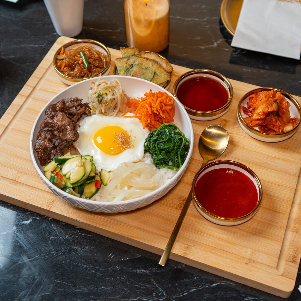 A colorful bibimbap bowl with beef, egg, and vegetables, served with kimchi and sauces on a wooden board.