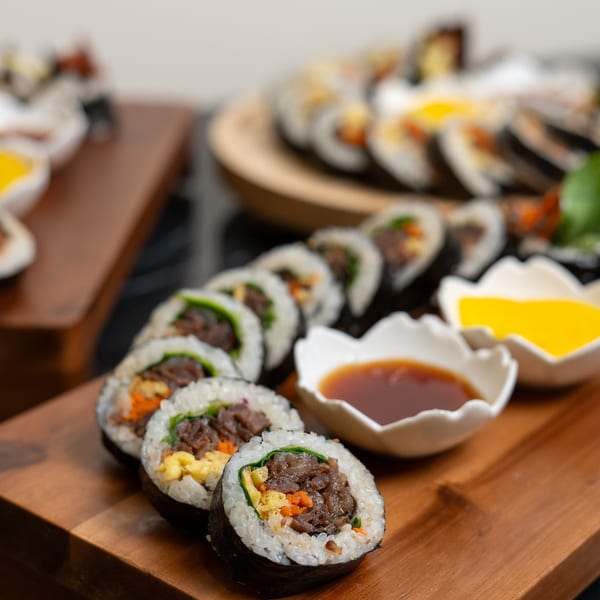 Rows of Korean gimbap rolls with beef, egg, and vegetables on a wooden board with dipping sauces.